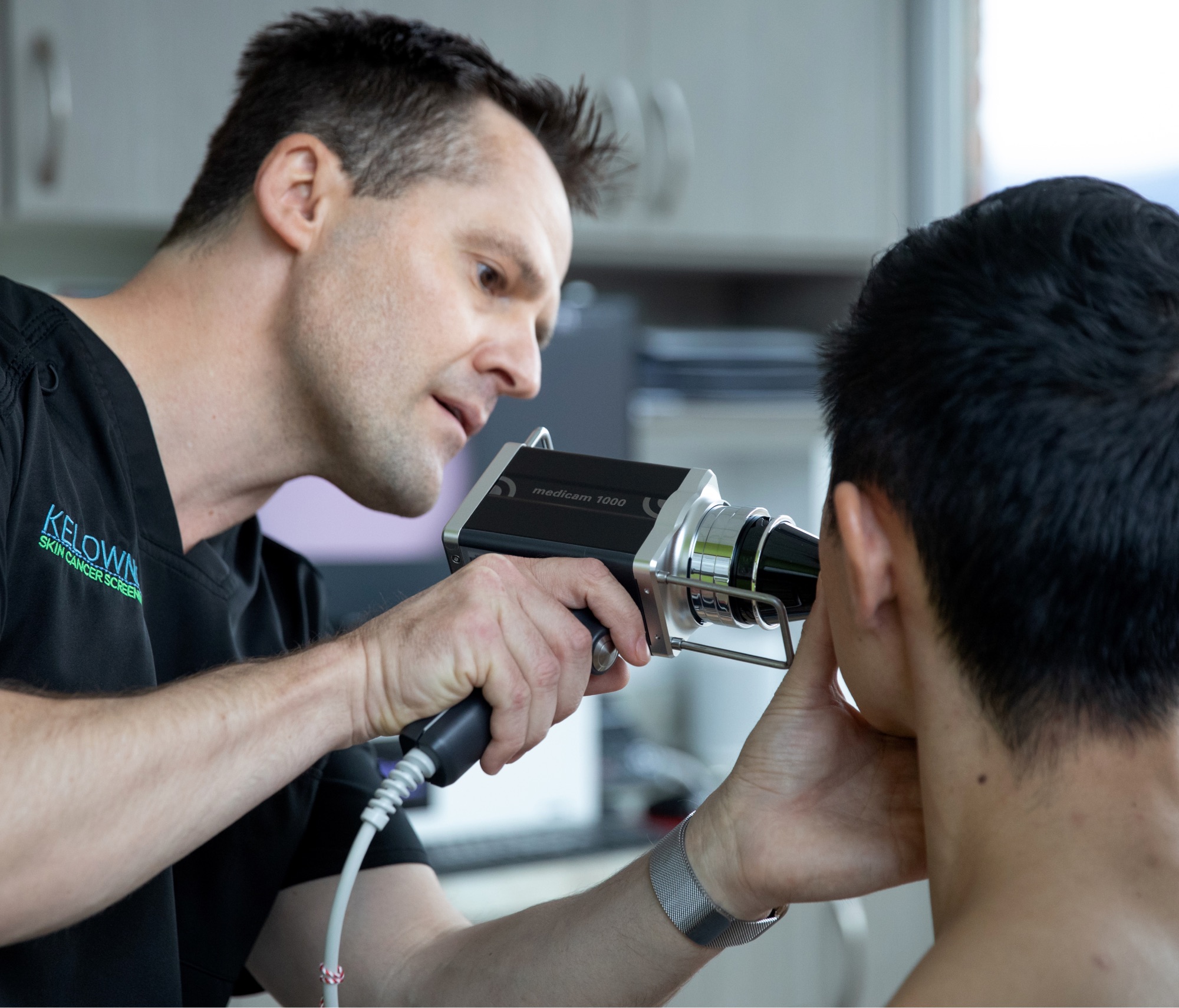 Dr Ben, a white man with short dark hair in scrubs, holding a visualizing tool to do a skin cancer treatment on a patient in his office. 