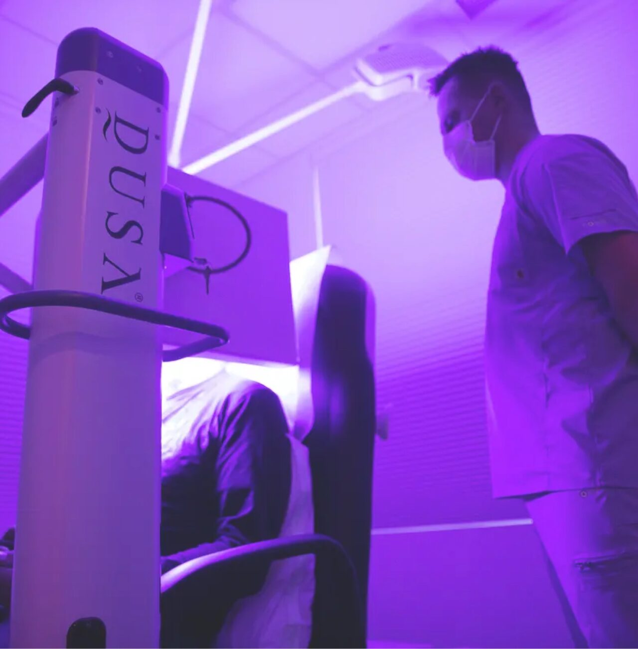 Dr Ben standing in the right side of the picture in scrubs and a mask beside a person sitting in a photodynamic therapy machine with DUSA written down the side. 